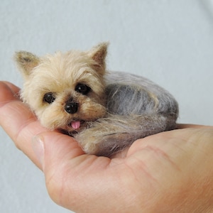 May include: A small, brown and gray Yorkshire Terrier dog figurine, lying on its side with its tongue sticking out, is held in a person's hand.