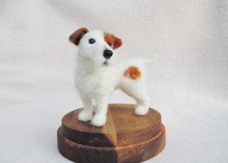 May include: A white and brown needle felted Jack Russell terrier dog figurine standing on a wooden base.