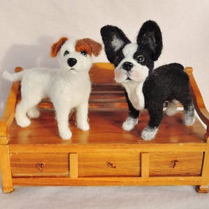 May include: Two felted wool toy dogs sitting on a wooden bench. The dog on the left is white with brown ears and the dog on the right is black and white with floppy ears.