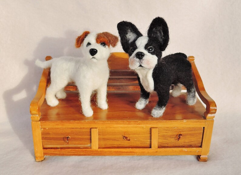 May include: Two felted wool toy dogs sitting on a wooden bench. The dog on the left is white with brown ears and the dog on the right is black and white with floppy ears.