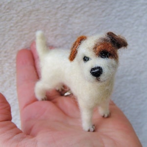 May include: A small, white, needle-felted Jack Russell Terrier dog with brown markings on its head. The dog is standing on a person's hand.