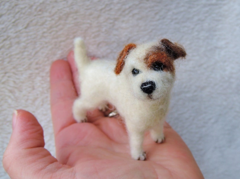 May include: A small, white, needle-felted Jack Russell Terrier dog with brown markings on its head. The dog is standing on a person's hand.