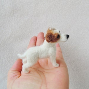 May include: A small, white, needle-felted Jack Russell terrier figurine with brown ears. The figurine is standing on a person's hand.