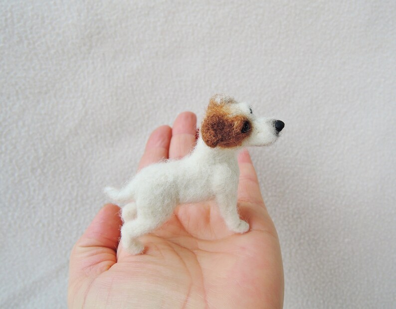 May include: A small, white, needle-felted Jack Russell terrier figurine with brown ears. The figurine is standing on a person's hand.