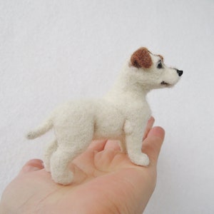 May include: A white felt Jack Russell Terrier dog with brown ears. The dog is standing on a person's hand.