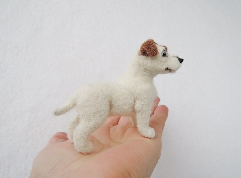 May include: A white felt Jack Russell Terrier dog with brown ears. The dog is standing on a person's hand.