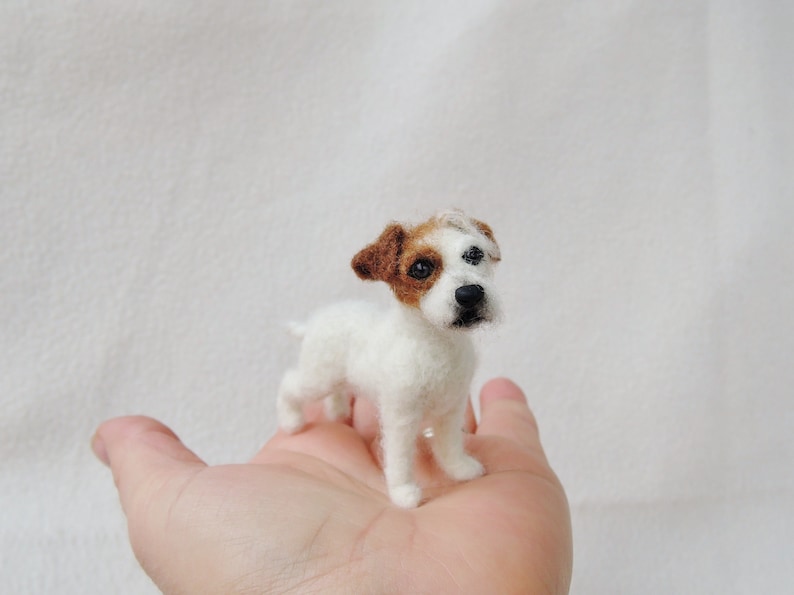 May include: A needle felted Jack Russell Terrier dog figurine. The dog is white with brown markings on its head and ears. It is standing on a person's hand.