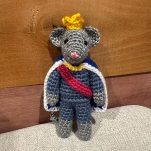 May include: A crocheted mouse doll featuring gray yarn, a yellow crown, and a blue cape with a white border. The mouse wears a blue tunic with a red sash across the chest and has a small pink nose. The figure is crafted with visible yarn stitches.