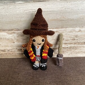 May include: A crocheted doll wearing a brown pointed hat, a black and grey robe, and a red and yellow scarf. The doll is holding a brown and grey broom.