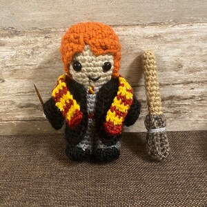May include: Crocheted amigurumi doll of a young boy with red hair, wearing a black robe and a yellow and red scarf. The doll is holding a small brown stick. The doll is standing on a brown surface.