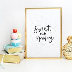 May include: A framed print with the words "Sweet as honey" in black script, a gold pineapple-shaped container, and a stack of books topped with decorative spheres and a cherry. The frame has a gold border.