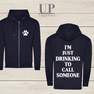 Könnte beinhalten: Marineblauer Kapuzenpullover mit Reißverschluss. Auf der Vorderseite befindet sich ein weißer Pfotenabdruck. Auf der Rückseite steht in Weiß: "I'M JUST DRINKING TO CALL SOMEONE". Der Hoodie hat eine Kapuze und einen durchgehenden Reißverschluss.