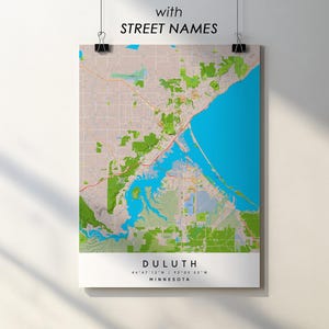 Pode incluir: Um cartaz com um mapa de Duluth, Minnesota, com nomes de ruas. O mapa detalha o layout da cidade, com massas de água azuis e áreas verdes. O texto "DULUTH" e as coordenadas estão na parte inferior. O cartaz é preso por clipes pretos.