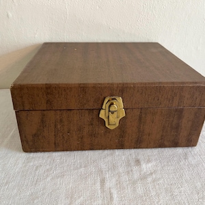 May include: A rectangular storage box with a brown wood grain finish and a gold-toned metal latch centered on the front panel. The box features a flat lid and a structured design, providing a simple and functional container for various small items.