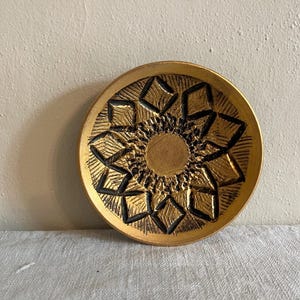 May include: A round, gold-colored decorative plate with a detailed geometric pattern in black. The plate features a central circular design surrounded by diamond and angular shapes. The plate is likely made of metal or ceramic.