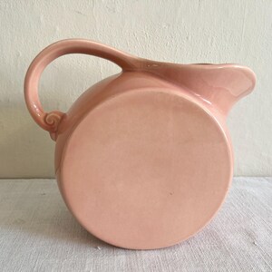 May include: A vintage, round, pink ceramic pitcher with a curved handle and a spout. The pitcher has a smooth, glossy finish and a simple, elegant design. The handle features a decorative swirl detail.