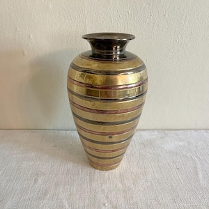 May include: A decorative brass vase with horizontal stripes in gold, copper, and dark gray. The vase has a rounded body and a narrow neck with a dark brown rim. The vase is displayed on a white surface.