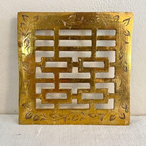 May include: A square gold-colored metal plaque featuring a central openwork design of the Chinese double happiness character. The border is decorated with an engraved leaf and vine pattern, creating a decorative frame around the central symbol.