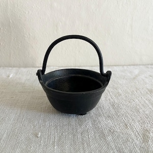 May include: A small, black cast iron cauldron with a rounded bowl and a curved handle. The handle is attached to the sides of the cauldron. The cauldron is sitting on a white, textured surface.