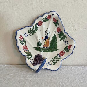May include: Leaf-shaped ceramic serving dish with a scalloped edge. The white dish features a hand-painted design of a woman in traditional clothing, flowers, and foliage. A cluster of purple grapes and a blue handle are at the base.