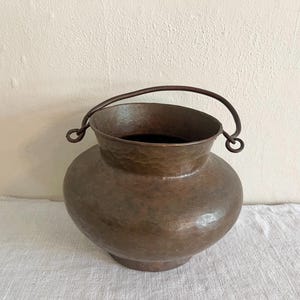 May include: A hammered copper bucket with a curved handle. The bucket has a rounded body and a wide opening. The handle is attached to the bucket with small, circular loops. The copper has a warm, reddish-brown color.