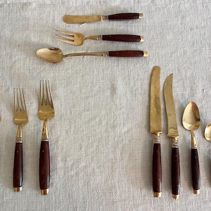 May include: A collection of vintage flatware with brass-colored heads and dark brown wooden handles. The set includes knives, forks, and spoons, arranged on a neutral-colored linen surface. The handles have a brass accent at the base.