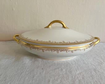 May include: A white porcelain serving dish with a lid, featuring gold trim and handles. The dish has a delicate floral pattern around the rim and on the lid, adding a touch of elegance to the design.