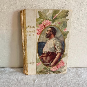 May include: Antique book titled "The Village Blacksmith" by Longfellow. The cover features an oval portrait of a man with a beard, surrounded by pink flowers and green leaves. The book has a worn, aged appearance.
