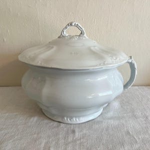 May include: A white ceramic chamber pot with a matching lid, featuring a curved handle on one side and a decorative knob on top. The vessel has a rounded, multi-faceted body with subtle embossed floral detailing along the rim and lid edges.