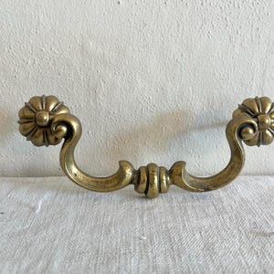 May include: A vintage brass drawer pull with a floral design on each end. The handle has a curved shape with a decorative center. The brass hardware is likely for a cabinet or dresser. The overall design is ornate and classic.