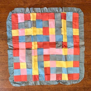 May include: A colorful, square quilt with a woven pattern of various colored satin ribbons. The ribbons are in shades of red, pink, yellow, blue, and gray. The quilt is trimmed with a ruffled, light green border.