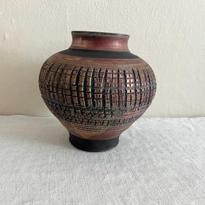May include: A handcrafted, textured ceramic vase with a unique, grid-like pattern. The vase features a dark red rim, a black band, and a brown body with a textured surface. The base is black, and the vase sits on a white surface.