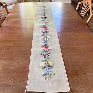 May include: A beige table runner with a red border features a repeating pattern of embroidered fruit, including pears, grapes, and apples. The runner is on a wooden table.