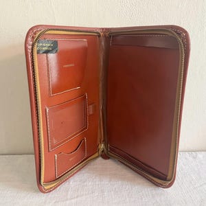 May include: A brown leather zippered portfolio featuring a top grain cowhide label, a clear identification window, multiple interior card slots, and a large document sleeve. The interior is lined with matching brown leather and white stitching.