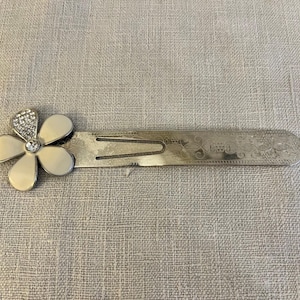 May include: Silver-toned metal bookmark with a flower design. The flower has four cream-colored petals and a center of clear rhinestones. The bookmark has a split end and decorative etching.