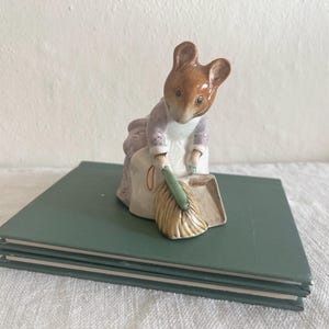 May include: A ceramic figurine of a mouse dressed in a lavender and white outfit, sweeping with a broom and dustpan. The figurine is set on a stack of green books. The mouse has brown fur and is bent over, focused on its task.