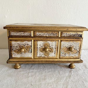 May include: A decorative wooden jewelry box with a distressed gold and white finish. The box features three small drawers with ornate gold hardware and is raised on small, rounded feet. The top and sides are also trimmed in gold.