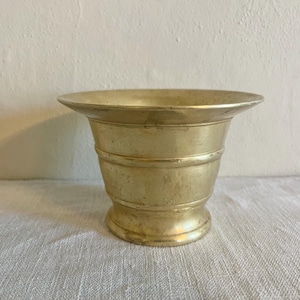 May include: A vintage brass mortar with a wide, flared rim and a cylindrical body. The mortar has horizontal bands around the middle and a small, rounded base. The brass has a warm, golden hue, suggesting age and use. This item is suitable for kitchen decor.