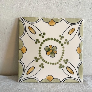 May include: Square ceramic tile with a floral design. The tile features a white background with a central flower and surrounding pattern in green, yellow, and black. The edges are outlined in green.