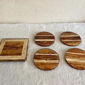 May include: A set of wooden coasters and a square tray. The coasters are round with alternating light and dark wood stripes. The tray has a square design with a light-colored border and a similar wood pattern.