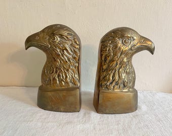 May include: A pair of brass eagle head bookends. Each bookend features a detailed eagle head with textured feathers and a curved beak. The bookends are mounted on rectangular bases, showcasing a vintage aesthetic.