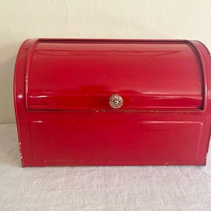 May include: A bright red metal bread box with a roll-top lid and a small, round, decorative silver-toned knob. The rectangular container features a smooth, glossy finish with visible signs of wear and paint chipping along the edges.