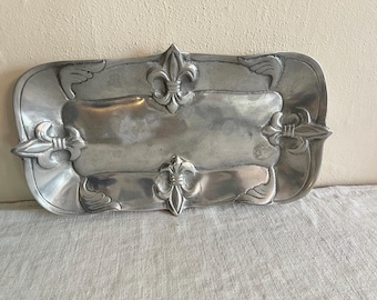 May include: A rectangular, silver-colored metal tray with a fleur-de-lis design. The tray has a raised border with decorative elements, including fleur-de-lis and stylized wing accents. The tray is approximately 12 inches long and 7 inches wide.