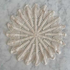 May include: Cream-colored, handmade crochet doily with a floral design. The circular doily features a central circle with intricate openwork patterns radiating outwards. The edges are scalloped, adding a decorative touch. This delicate piece is suitable for home decor.