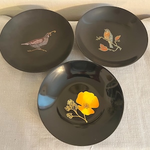 May include: Three black decorative plates with bird and floral designs. One plate features a bird with a worm, another a robin on a branch, and the third a yellow flower. The plates are round and have a glossy finish.