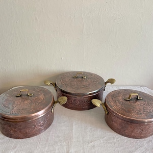 May include: Three copper cooking pots with lids and decorative handles. The pots have an aged, antique appearance with intricate floral patterns etched into the metal. The handles are a contrasting gold color.