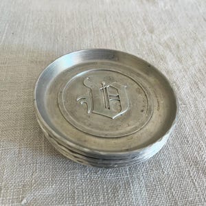 May include: A circular silver-toned metal coaster featuring a raised, embossed gothic-style letter D in the center. The coaster has a smooth, slightly recessed interior surface and a textured, hammered-style edge around the outer rim.