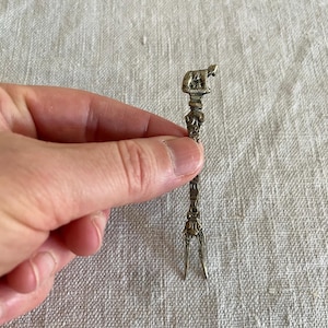 May include: A silver-toned, decorative fork with a detailed design. The fork features a small animal figure at the top and intricate patterns along the handle. The tines are split at the bottom. The fork is held in a hand.