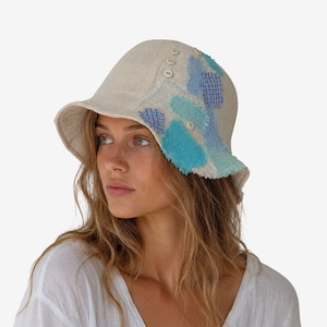 Può includere: Un cappello a secchiello beige caratterizzato da un design a toppe testurizzate con bordi sfrangiati in tonalità blu e turchese. Il cappello è arricchito da piccoli bottoni rotondi di colore chiaro sul lato, che aggiungono un dettaglio sottile.