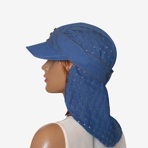 May include: A blue denim cap featuring a stiff front brim and a long, integrated neck flap that extends down the back. The entire surface of the cap and the neck flap is covered in a repeating pattern of small, circular, metal-rimmed eyelet holes.